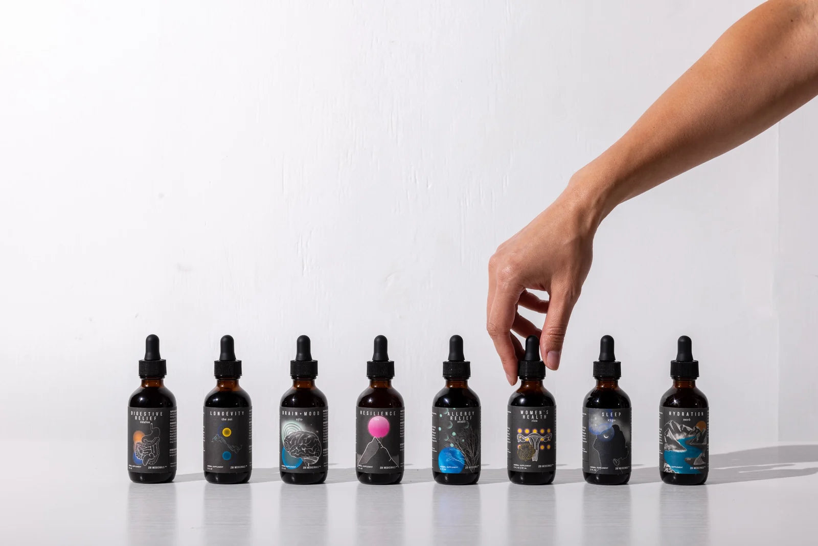 Eight Zoi formulas in 4 oz amber dropper bottles, lined up on white, with a hand reaching for one